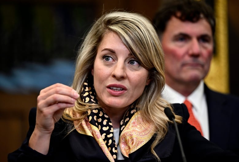 A woman wearing a shawl speaks at a microphone in outside of the House of Commons. A man in a suit and tie stands in the background.