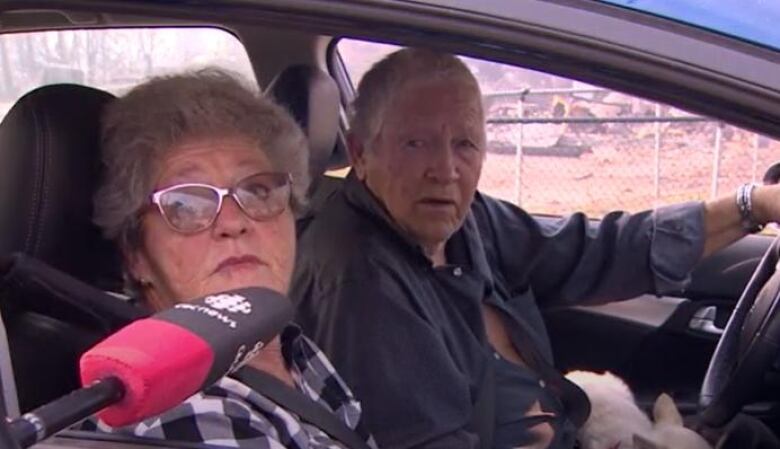 An older man and a woman sit in a vehicle, with someone holding a CBC microphone at the passenger window.