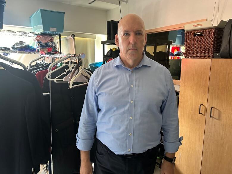 A man looks seriously at the camera in a room with shelves full of clothes to be donated. 