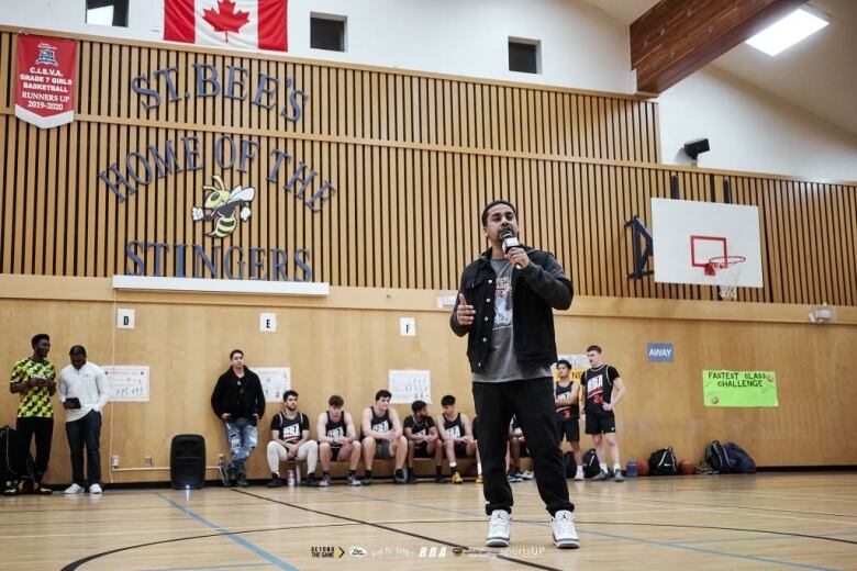 A brown man speaks into a mic while on a basketball court.