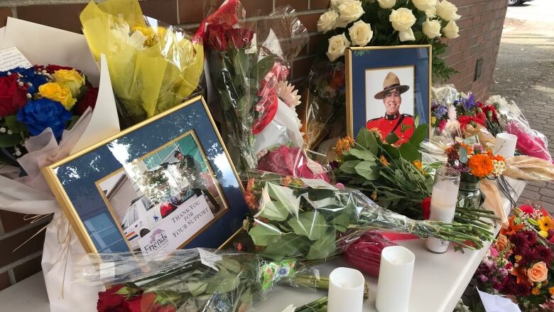 Two photos, each framed by a navy blue border, are pictured on a while table surrounded by bouquets and unlit candles. One of the photos, on the right, shows a man in a red uniform and brown hat. The other photo shows the man pictured in a black police uniform, taking groceries out of a car's boot. A piece of paper on that image reads, 'Thank you for your support.'