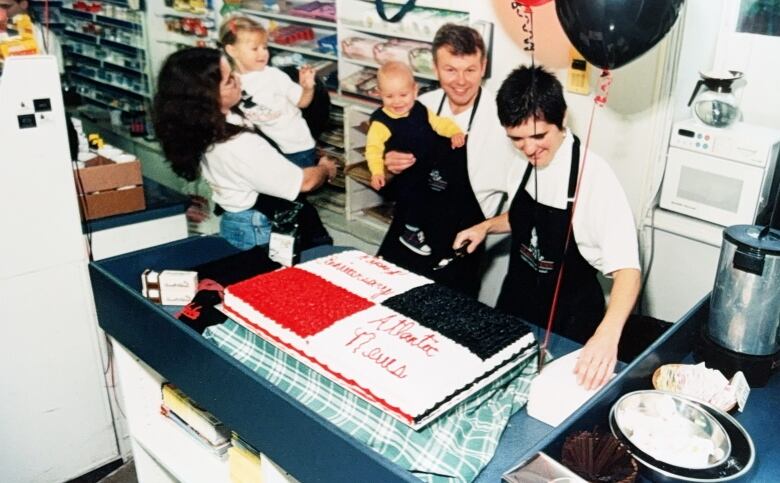 A man holding a baby and a women smile for the camera in front of a large cake. Another woman holding a child can be seen on the far left. 