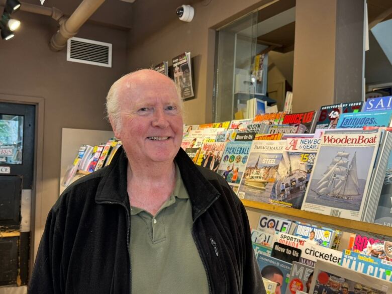 A man, with white hair, and a green polo, stand next to a rack of magazines.