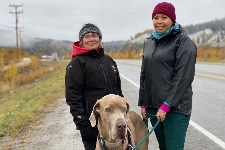 Two women standing with a dog on the side of the road