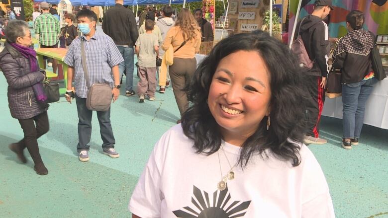 A person wearing a white tshirt speaks to camera while surrounded by a crowded market of stalls and vendors