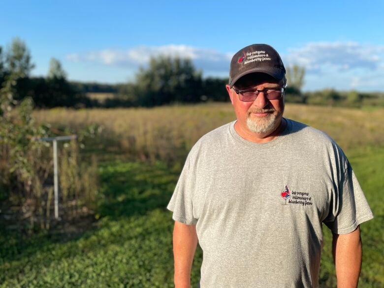 A man wears a shirt with the name of his farm on it as he stands near a field.