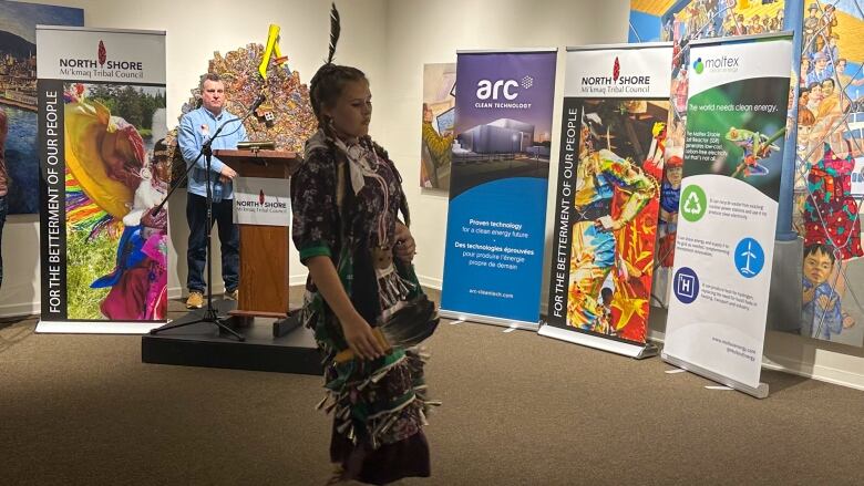 Man at a podium watches a young woman in traditional jingle dance attire. 