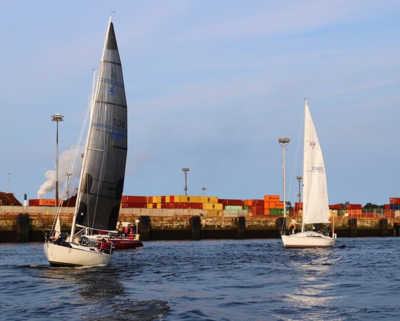 Two sailboats cruise past shipping containers stacked on the dock of west Saint John.