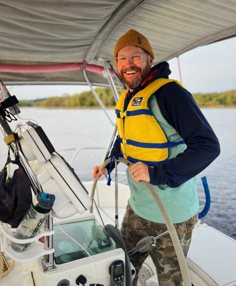 A smiling bearded man steers a sailboat past the city of Saint John