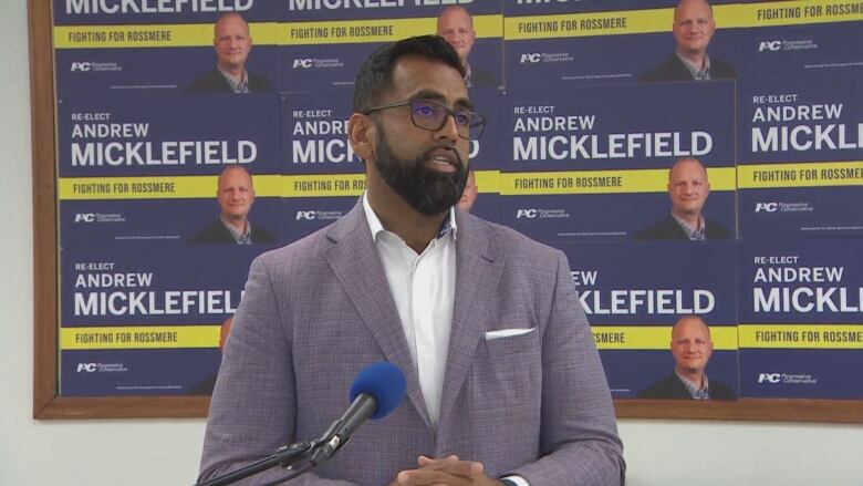 A man in a suit and glasses speaks at a microphone in front of a wall of campaign signs.