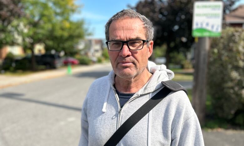 A man with glasses and grey sweater stands on a street.