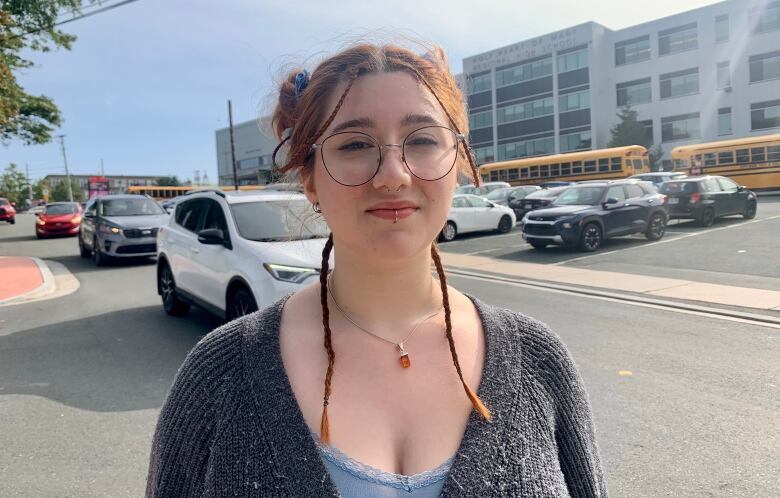 A young woman stands in front of a high school wearing glasses and a grey cardigan.