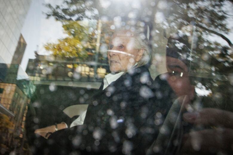 A man with white hair is seen sitting handcuffed inside a car.