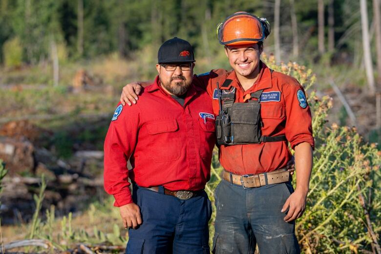 Two men wearing red shirts and wildfire management gear pose with their arms around each other