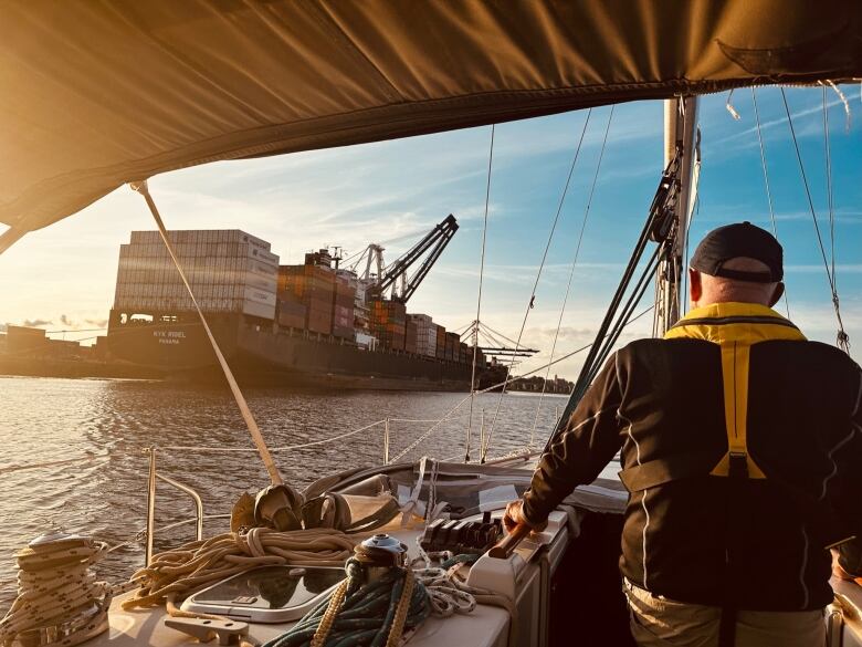 A man surveys a large container ship from the deck of a sailboat in solden sunset light.