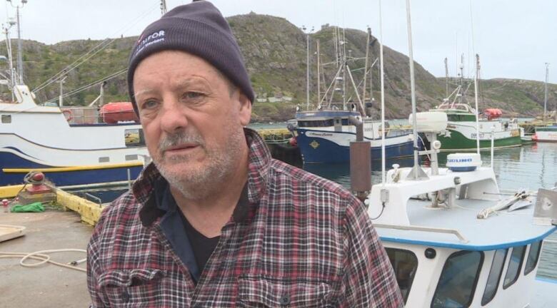 A man in a dark tuque and plaid shirt stands on a wharf in front of small fishing boats.