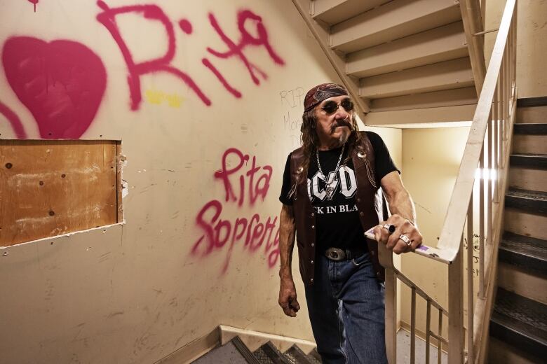 A man in a leather jacket stands in the hallway of an apartment building with graffiti in the background.