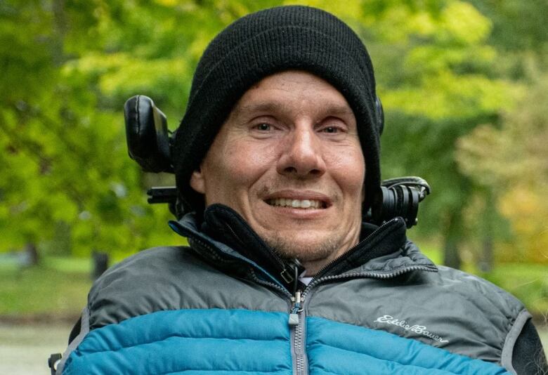 A man wearing a black toque and blue puffer vests smiles at the camera.