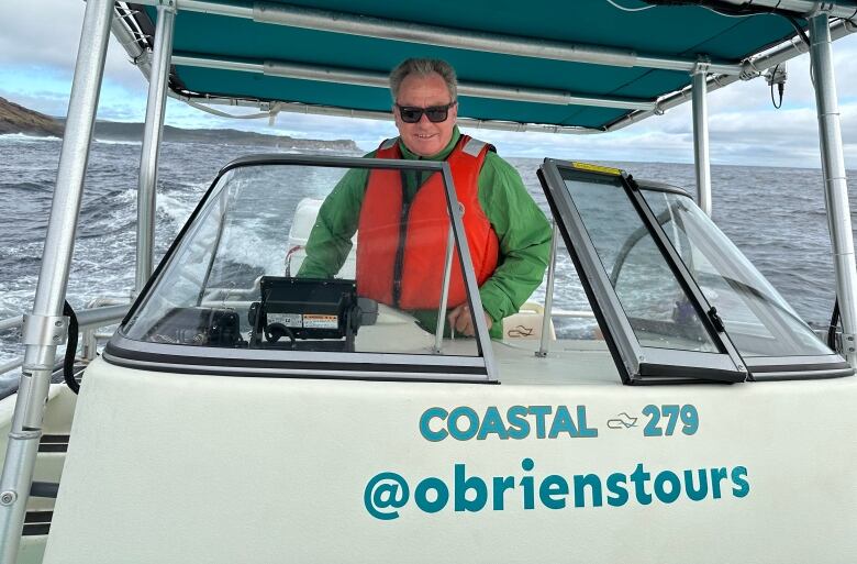 A man in a green jacket and orange life-jacket pilots a small boat.