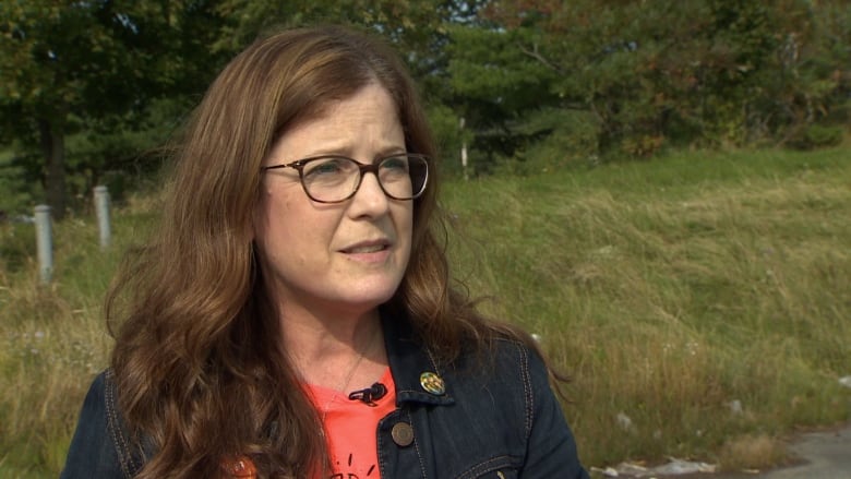 A woman with brown hair and glasses looks off to the side. She wears a jean jacket and orange shirt. Behind her is grass and trees. 