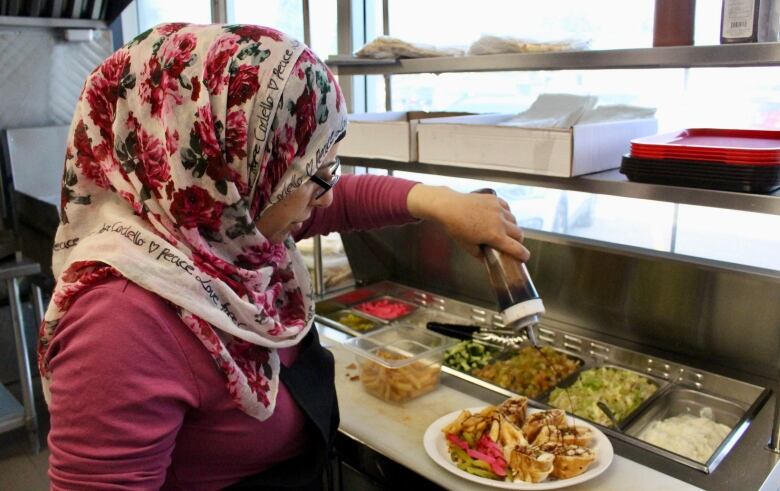 A woman in a commercial kitchen drizzles sauce on a plate of cut up pieces of a chicken wrap. There are metal containers of vegetable toppings in front of her.