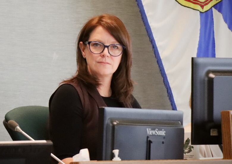 A woman with auburn hair and large glasses sits at a computer monitor with a Nova Scotia flag in the background.