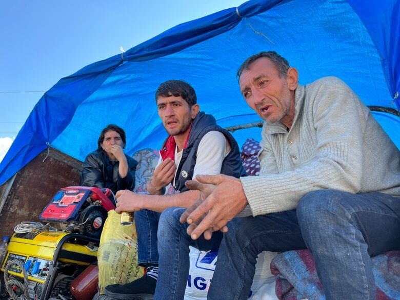 Two men sit in the back of a truck that is covered with a tarp and piled with belongings. A woman sits behind them.