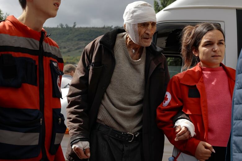 An older man with white bandages on his head walks with the help of two aid workers who flank him.