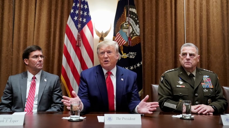 A man in a suit and tie gestures with his hands while speaking. He is seated at a conference table, with a man in a suit and tie to his right, and a man in a military uniform to his left.