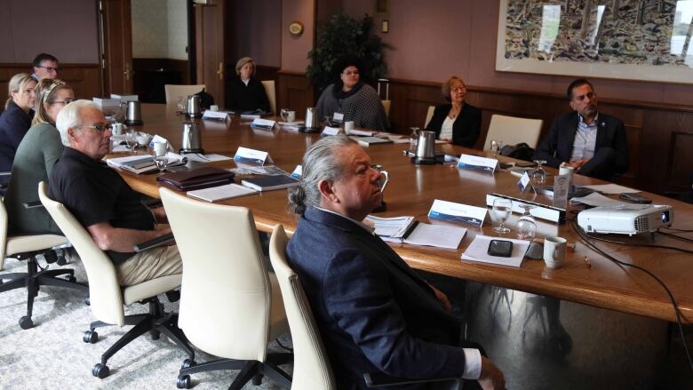 A group of people seated around an oval shaped table look to the right of the image at something out of frame