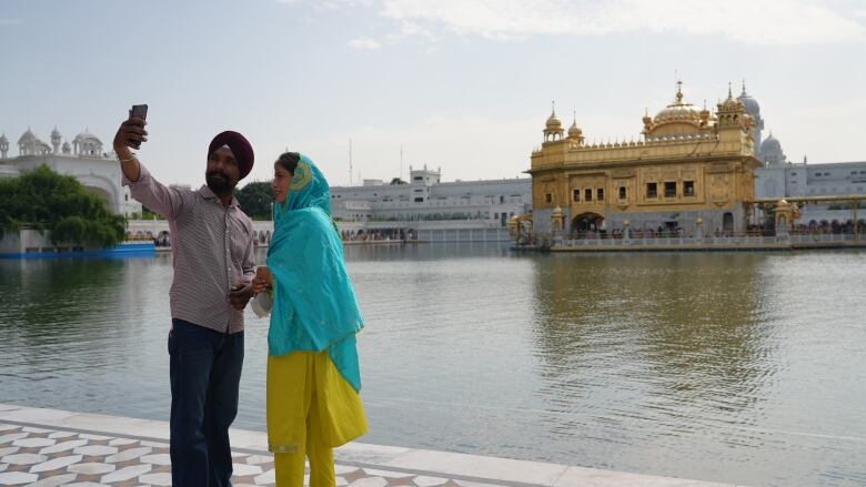 Two people take a selfie beside a body of water in front of a golden temple.