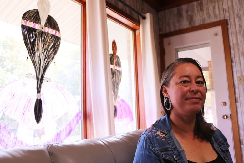 A woman in a jean jacket sits on a couch in front of windows on which Indigenous women with beads in their hair were painted.