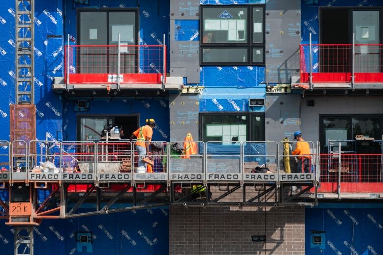 Two construction workers on a lift work on a building covered in blue construction wrap.