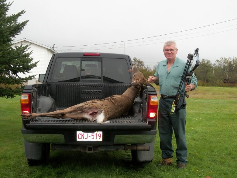 A man with gray hair, a green shirt and pants and a crossbow, stands next to the open tailgate of a pick-up truck, parked on grass, containing a dead deer.