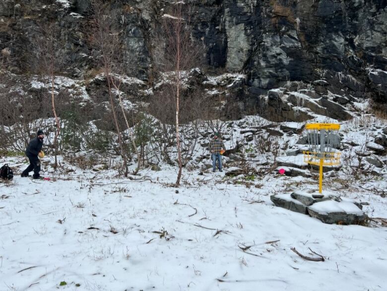 Two disc golfers prepare to play a hole of disc golf. There's snow on the ground, which is covering some rocky terrain.