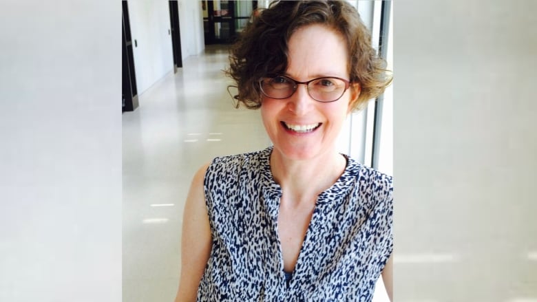 A woman with short, dark curly hair and burgundy-rimmed glasses is pictured in a sleeveless, black and white textured blouse, smiling at the camera. 