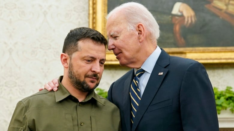 A taller grey-haired man in a suit, puts his hand on the shoulder of a shorter, bearded man in a green button-up shirt. 