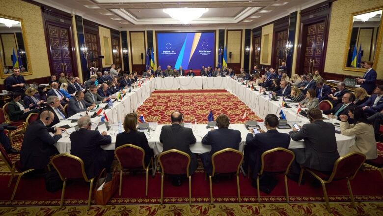 Dozens of people, mostly men in suits, sit around tables set up as a large rectangle in a well-appointed, carpeted room.