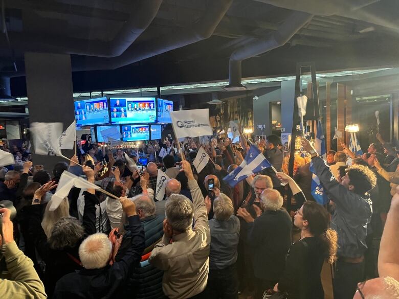 People raise flags and look at screens in a crowded room. 