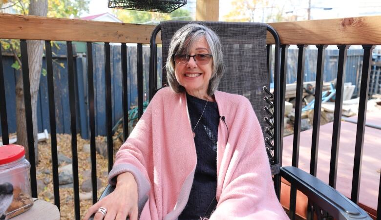 A woman wearing glasses sits outside in a chair. She is wearing a pink sweater over a black shirt. 