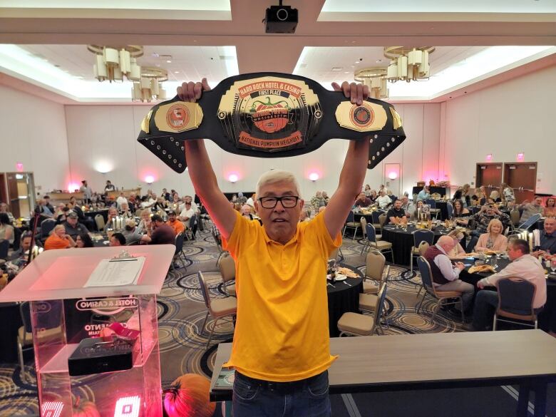 A man stands in a hall holding up a giant trophy belt. 