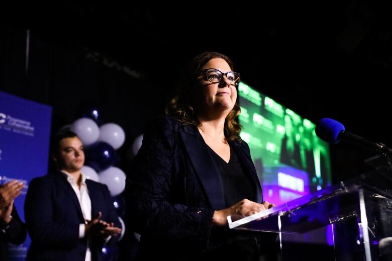 A woman with shoulder-length hair and glasses speaks to an audience.