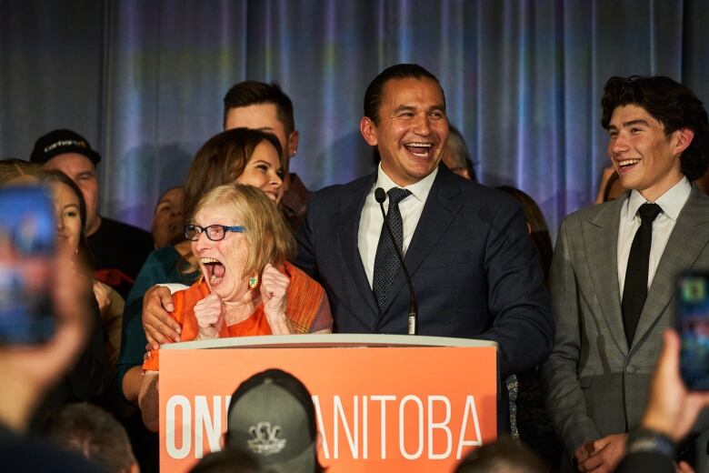 A smiling man in ablue suit puts his arm around a woman in an orange dress as they both stand behind a podium.