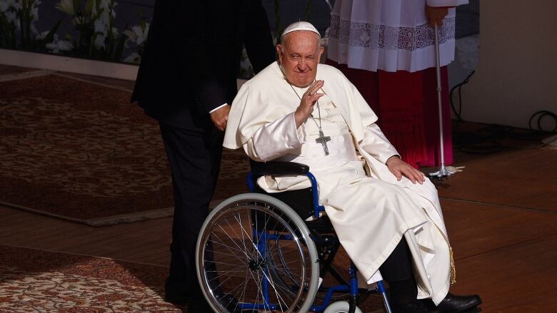 The Pope is shown in a wheelchair wearing white papal garments and cap.