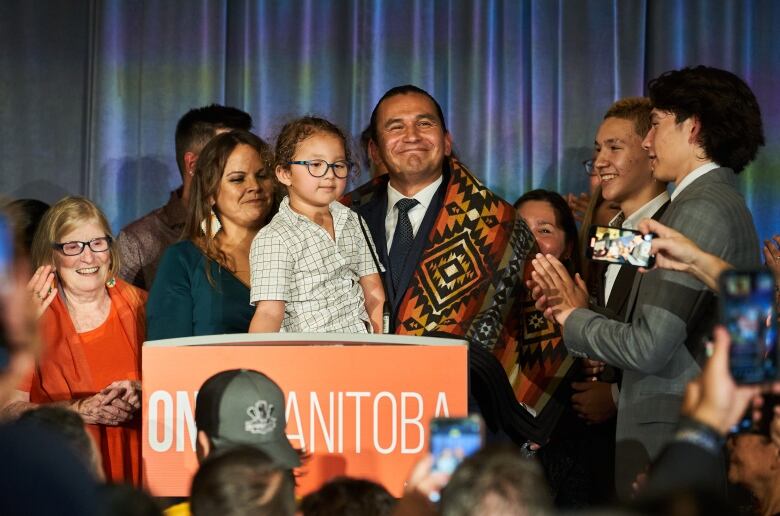 Manitoba NDP leader Wab Kinew stands with his family a smiles at a podium before giving his victory speech.