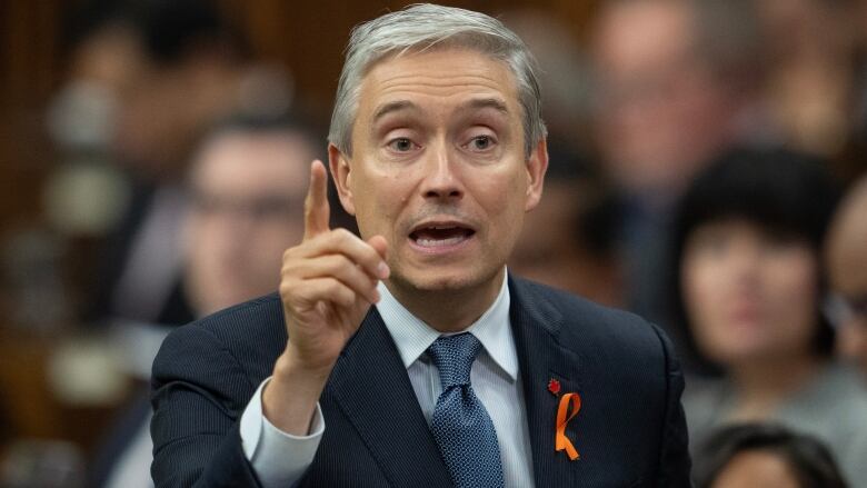 A man in a suit faces the camera and raises one index finger.