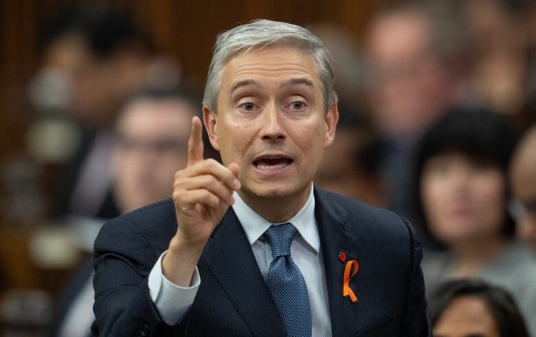 A man in a suit faces the camera and raises one index finger.