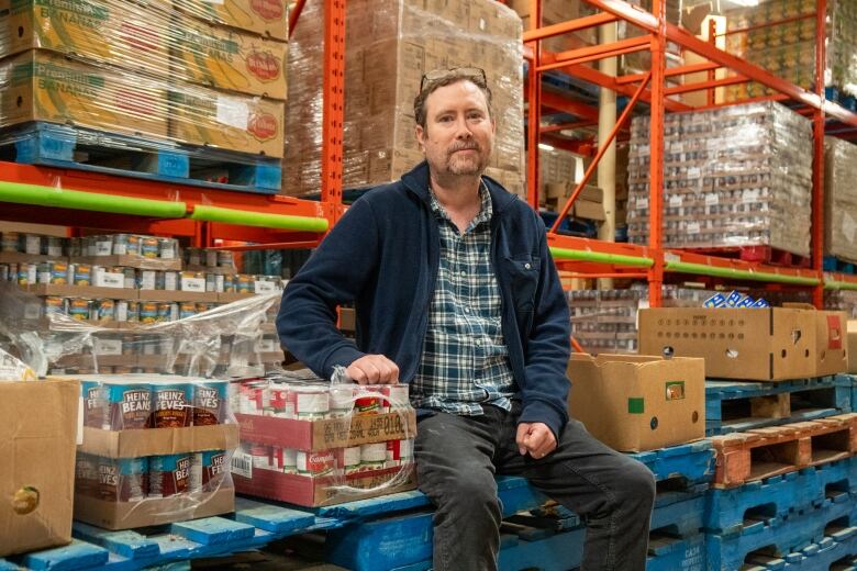 A man sits on a stack of crates beside packages of beans in a warehouse.