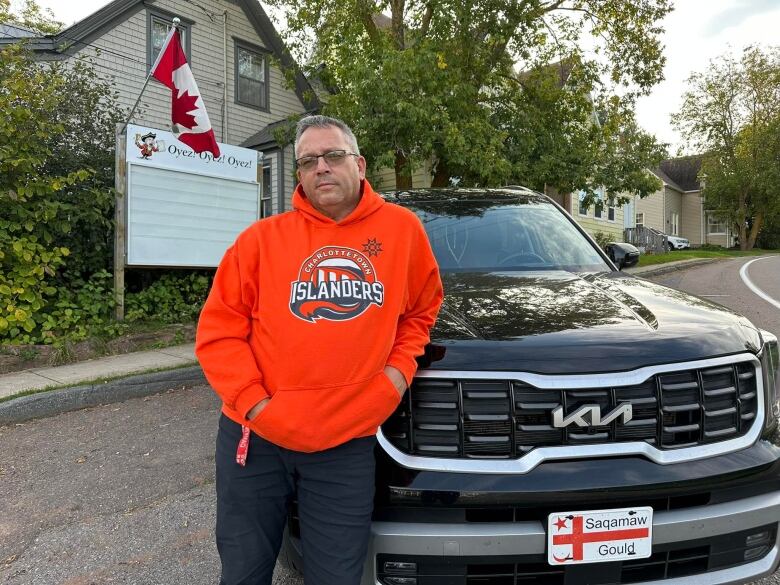 Man in orange Charlottetown Islanders hoodie leans on hood of truck with empty sign and Canada flag visible in background.