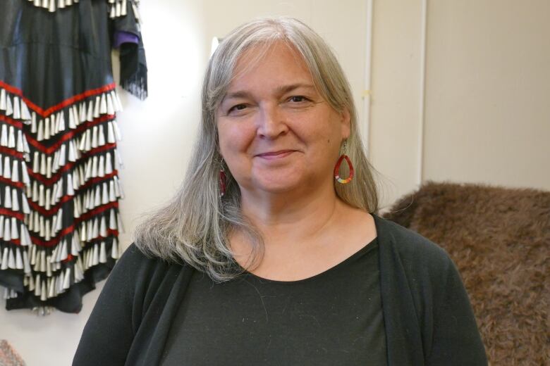 A woman with shoulder-length grey hair sits in front of a First Nations jingle dress and a bison rug hanging behind her.